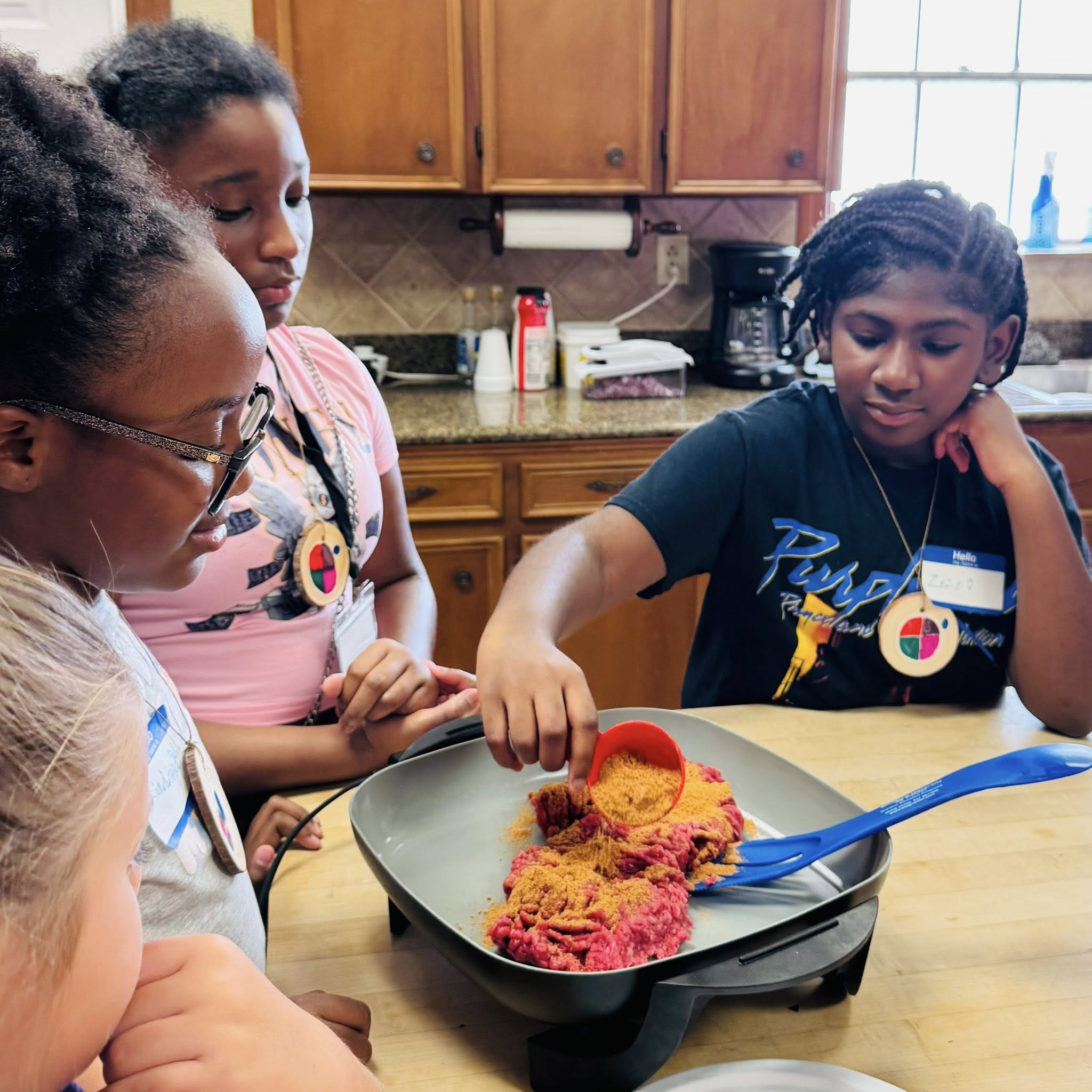 Two girls watching another girl pour seasoning over meat in a skillet.